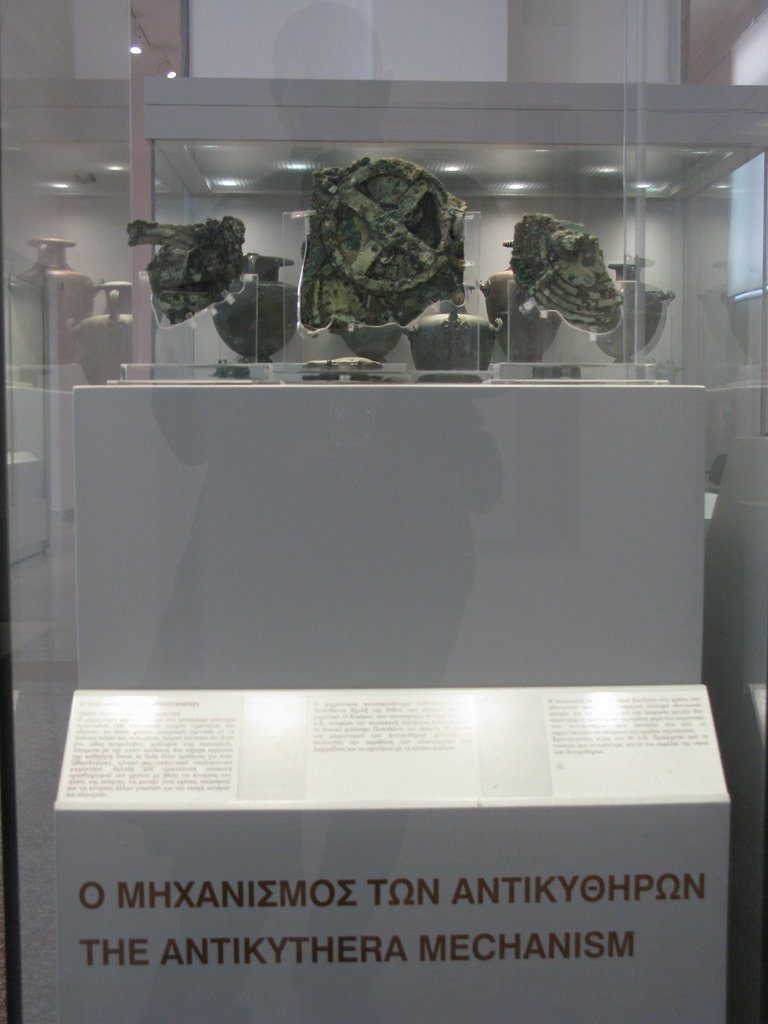 Photographic image of The Antikythera Mechanism exhibit at the National Archeological Museum of Athens. Grecian urns are on display in the background.
