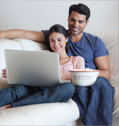 Photo of a couple on a couch, looking at a laptop screen and holding a bowl of popcorn