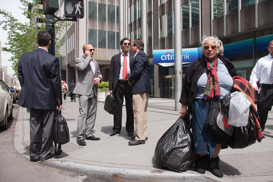 Photo shows a homeless elderly woman and a few young men in executive formal attire standing in front of office buildings on the road side.