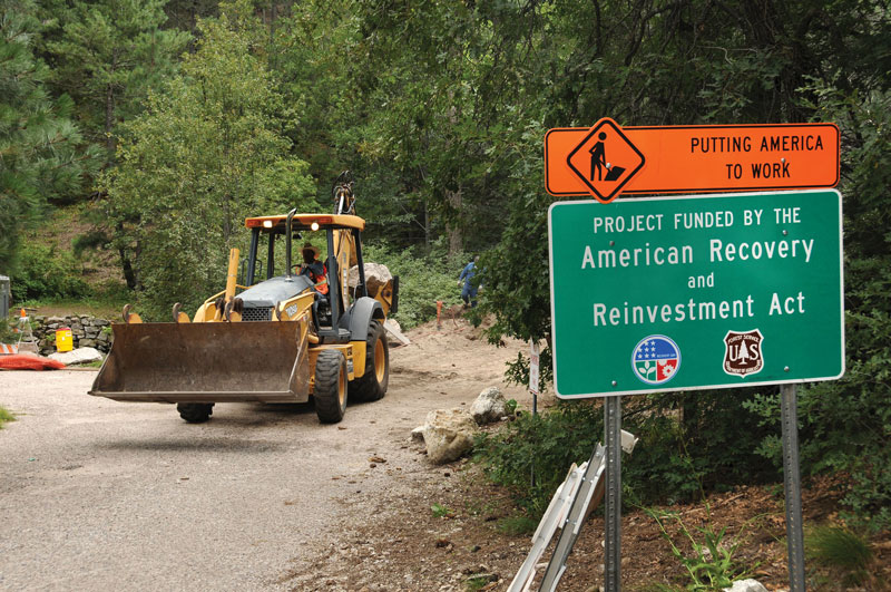 Photo shows an earth moving machine near a board which reads, “Project funded by American recovery and reinvestment act”.