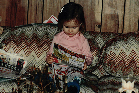A photograph of young Lindy Ryan reading newspaper, while seated on a couch.