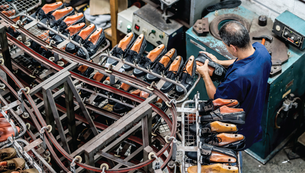 A man in a factory fits a leather shoe onto a form amongst an assembly line of shoe forms.