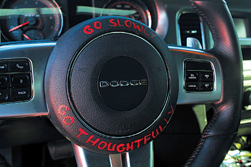 A color photograph depicting steering wheel of Dodge, where the upper and lower parts of central panel reads “Go Slowly” and “Go Thoughtfully”.