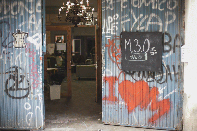 A photograph of a studio/office with open steel doors. The doors are painted with text and symbols and a black-board is hung that reads M 3.0. Inside the room a chandelier is present followed by another room where some things are kept.