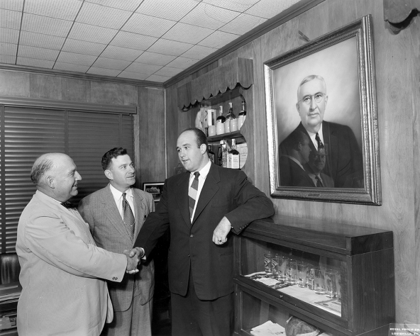 Photograph depicting Booker shaking hands with Uncle Jere while his cousin Carl is looking at him. Behind Booker on the wall is a portrait of Jim Beam, and on the left of the wall are shelves with some bottles. Below the portrait is a show-case containing glasses.