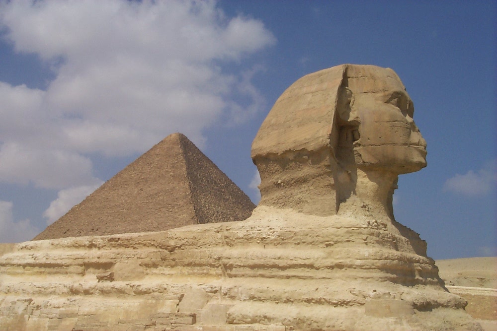 Image of The Great Sphinx at Giza with upper half of the Great Pyramid in the background. The surface of the Sphinx appears well worn by the ravages of time. The image is evocative of ancient Egypt.