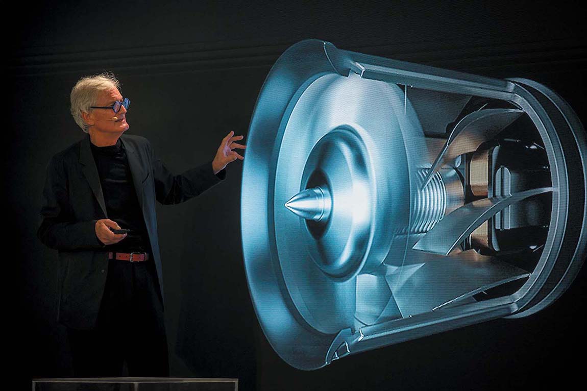 James Dyson standing in front of a cylindrical shaped object with spiral blades attached to its body and a conical tip at the center.