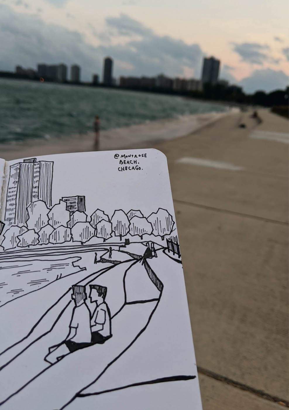 A sketchbook drawing of Montrose Beach in Chicago, held up against the real-life scene of the beach and skyline at sunset.