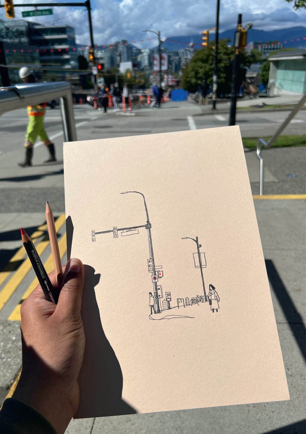 A hand-drawn sketch of a street scene in Vancouver, featuring a traffic light, pedestrians, and minimalistic details, held against the real-life background of the same scene.