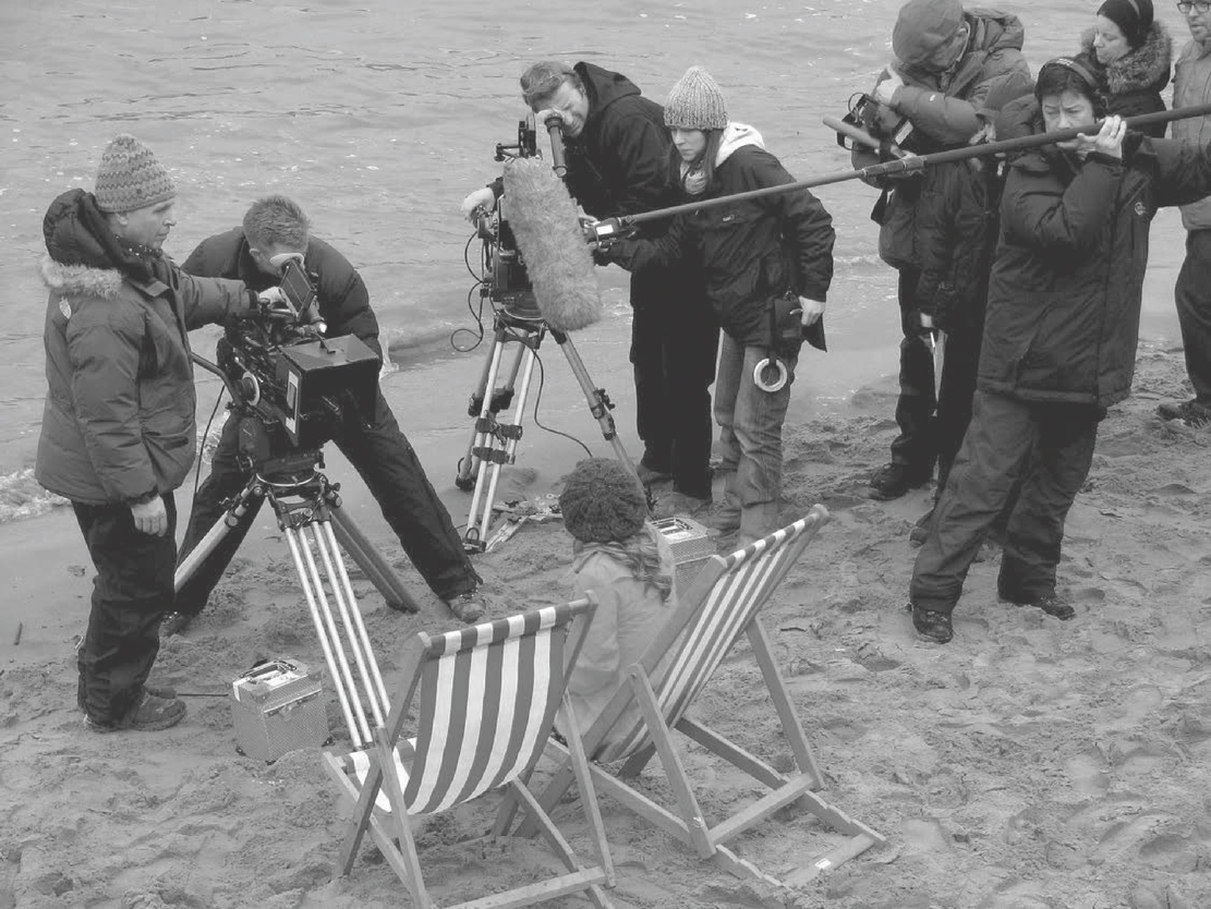 A photo presents a camera operator, a boom operator holding a shotgun microphone, and crew members surrounding an actor seated in a deck chair.