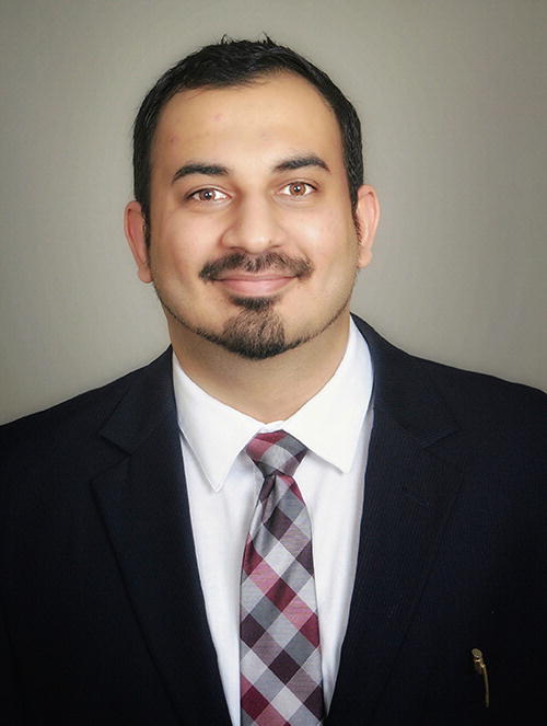 A person in a formal suit and tie is smiling at the camera. The background is plain, and the person has short hair and a beard. The tie features a checkered pattern with shades of red, gray, and white. A pen is visible in the suit pocket.
