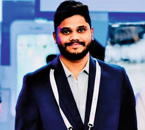 A person in a suit is standing indoors, smiling at the camera. They are wearing a dark blazer over a light shirt, with a lanyard around their neck. The background features blurred elements, including a large screen and other indistinct objects, suggesting a professional or event setting.