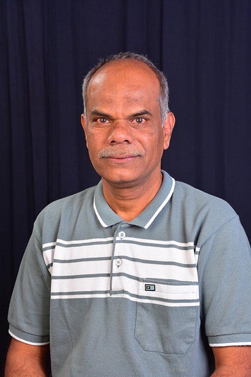 A person is standing against a dark blue curtain backdrop, wearing a light blue polo shirt with white horizontal stripes. The person is looking directly at the camera with a neutral expression.