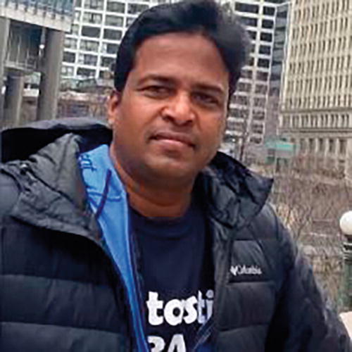 A person wearing a black jacket and a dark shirt with text stands outdoors in an urban setting. The background features tall buildings and a cloudy sky. The person appears to be looking at the camera.