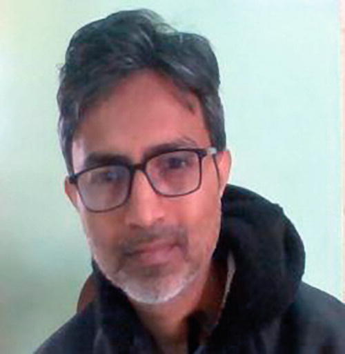 A person with short hair and glasses is looking at the camera with a neutral expression. They are wearing a dark hoodie. The background is a plain light green wall.