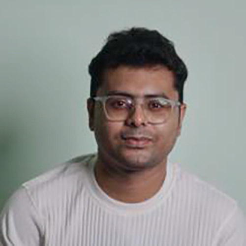 A person wearing glasses and a white ribbed shirt is sitting against a plain light green background. The individual is looking directly at the camera with a neutral expression.