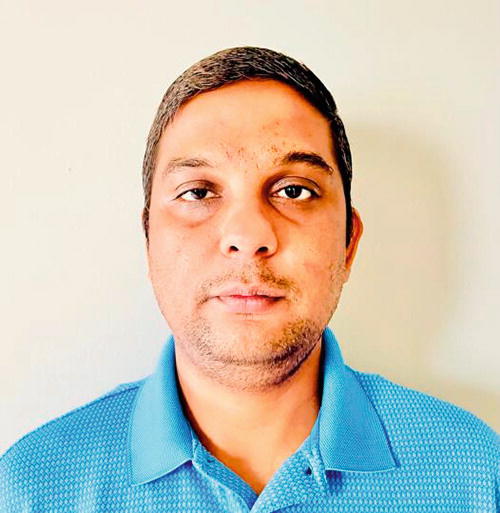A person wearing a blue polo shirt stands against a plain light-colored background. The individual is looking directly at the camera with a neutral expression.