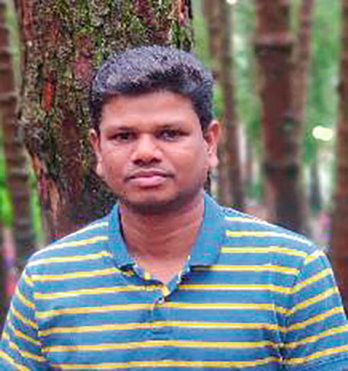 A person standing outdoors in a forested area, wearing a blue and yellow striped polo shirt. The background features tall trees with textured bark, creating a natural setting. The person has a neutral expression.