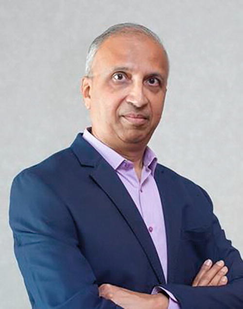 A person in a blue suit and light purple shirt stands with arms crossed against a plain background. The individual has a neutral expression and is looking directly at the camera.
