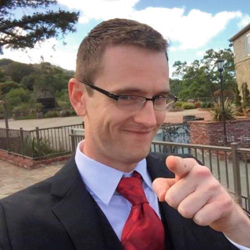 A person in a suit and red tie is smiling and pointing towards the camera. They are wearing glasses and standing outdoors on a patio with a scenic background of trees and a building. The sky is partly cloudy. The expression is friendly and engaging.