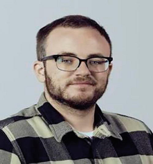 A person with short hair and glasses is wearing a plaid shirt. They are looking at the camera with a neutral expression. The background is plain and light-colored.
