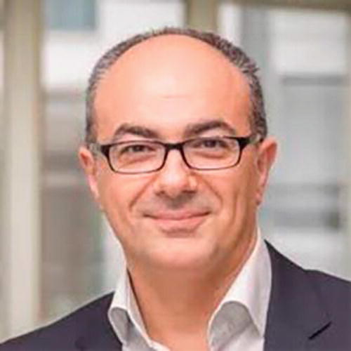 A person wearing glasses and a suit is smiling in an indoor setting with blurred background elements. The image conveys a professional and approachable demeanor.