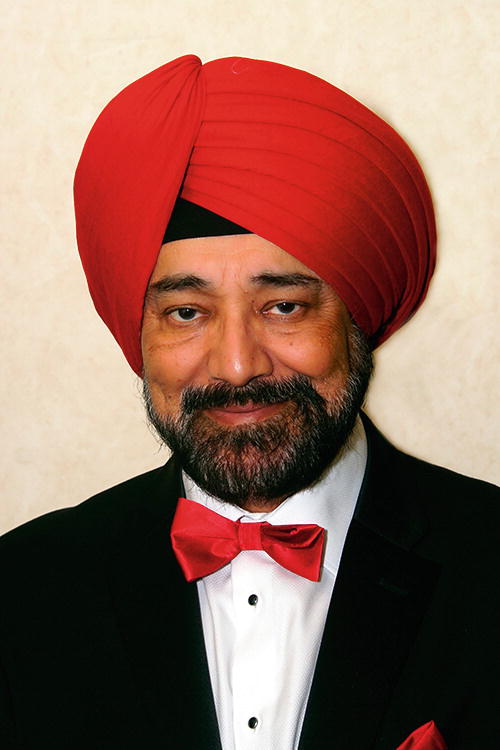 A person wearing a red turban and a black suit with a white shirt and red bow tie, smiling at the camera. The background is a plain, light-colored wall.