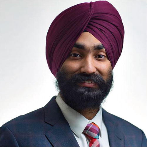 A person wearing a maroon turban and a dark suit with a striped tie is smiling. The background is plain and light-colored, emphasizing the individual. The image conveys a professional and approachable demeanor.