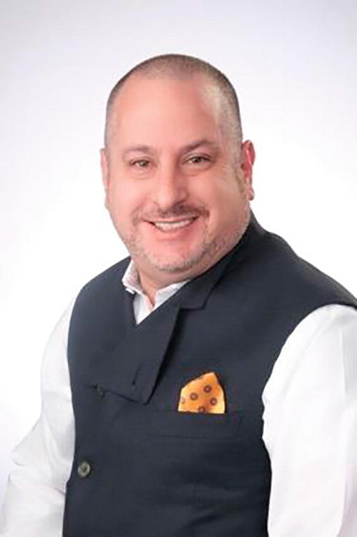 A person smiling, wearing a dark vest over a white shirt, with a patterned pocket square. The background is plain and light-colored.