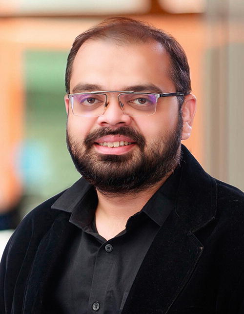 A person wearing glasses and a black jacket is smiling in a professional setting. The background is softly blurred with warm tones, suggesting an indoor environment.