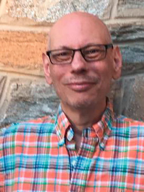 A person wearing glasses and a colorful plaid shirt stands in front of a stone wall. The person is smiling, and the background features textured stonework in shades of beige and gray.