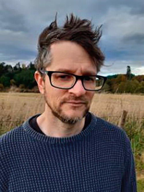 A person wearing glasses and a sweater stands in a field with tall grass. The background features a cloudy sky and a line of trees, suggesting an outdoor, rural setting. The person appears to be looking at the camera.