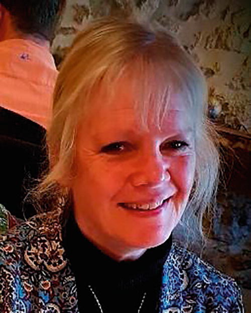 A person smiling in a warmly lit indoor setting with a stone wall in the background. The individual is wearing a patterned garment and a dark top. The atmosphere appears cozy and relaxed.