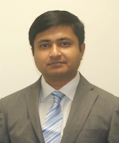 A person wearing a gray suit, white shirt, and blue striped tie, standing against a plain light-colored background.