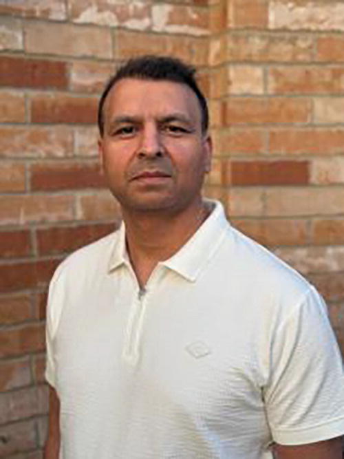 A person standing in front of a brick wall, wearing a light-colored polo shirt. The expression is neutral, and the setting appears to be outdoors.