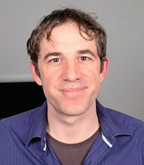 A person is smiling at the camera, wearing a blue shirt over a dark undershirt. The background is plain and neutral.