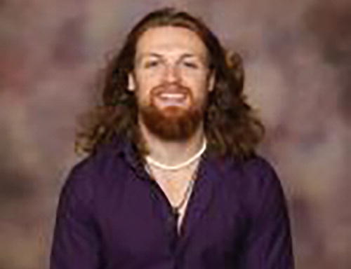 A person is seated against a blurred, neutral-toned background. They are wearing a dark purple shirt and a necklace. The person is smiling and looking towards the camera.