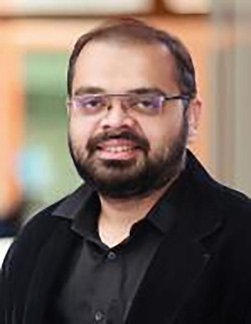 A person wearing glasses and a black jacket is smiling slightly. The background is softly blurred, featuring warm tones.