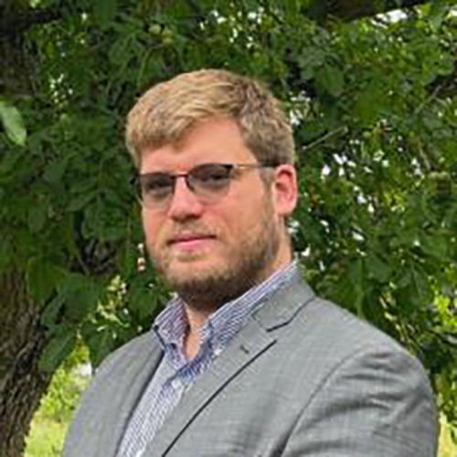 A person wearing glasses and a suit stands outdoors in front of a leafy tree. The background is lush and green, suggesting a natural setting.
