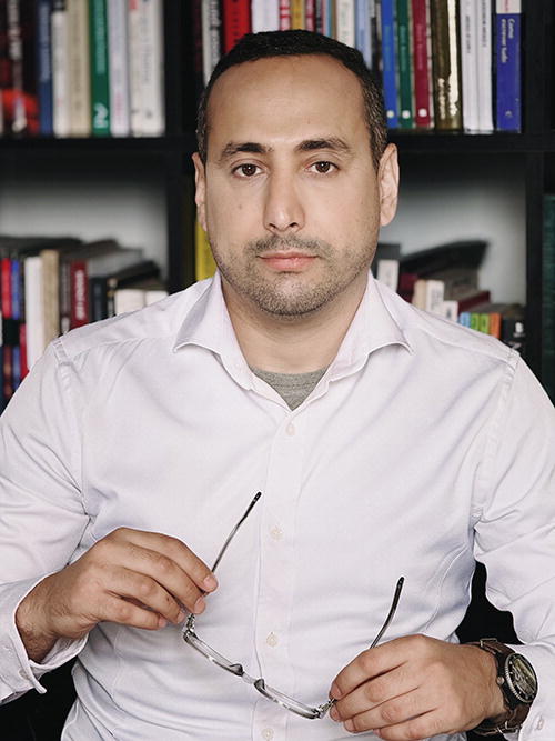 A person wearing a white shirt is holding a pair of glasses in front of a bookshelf filled with various books. The individual is looking directly at the camera with a neutral expression. The background features a variety of book spines, suggesting a setting related to study or work.