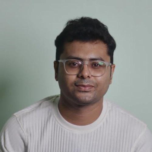 A person wearing glasses and a light-colored sweater is seated against a plain background. The individual is looking directly at the camera with a neutral expression.