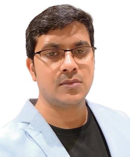 A person wearing glasses and a light-colored blazer over a dark shirt, looking towards the camera with a neutral expression. The background is plain white.