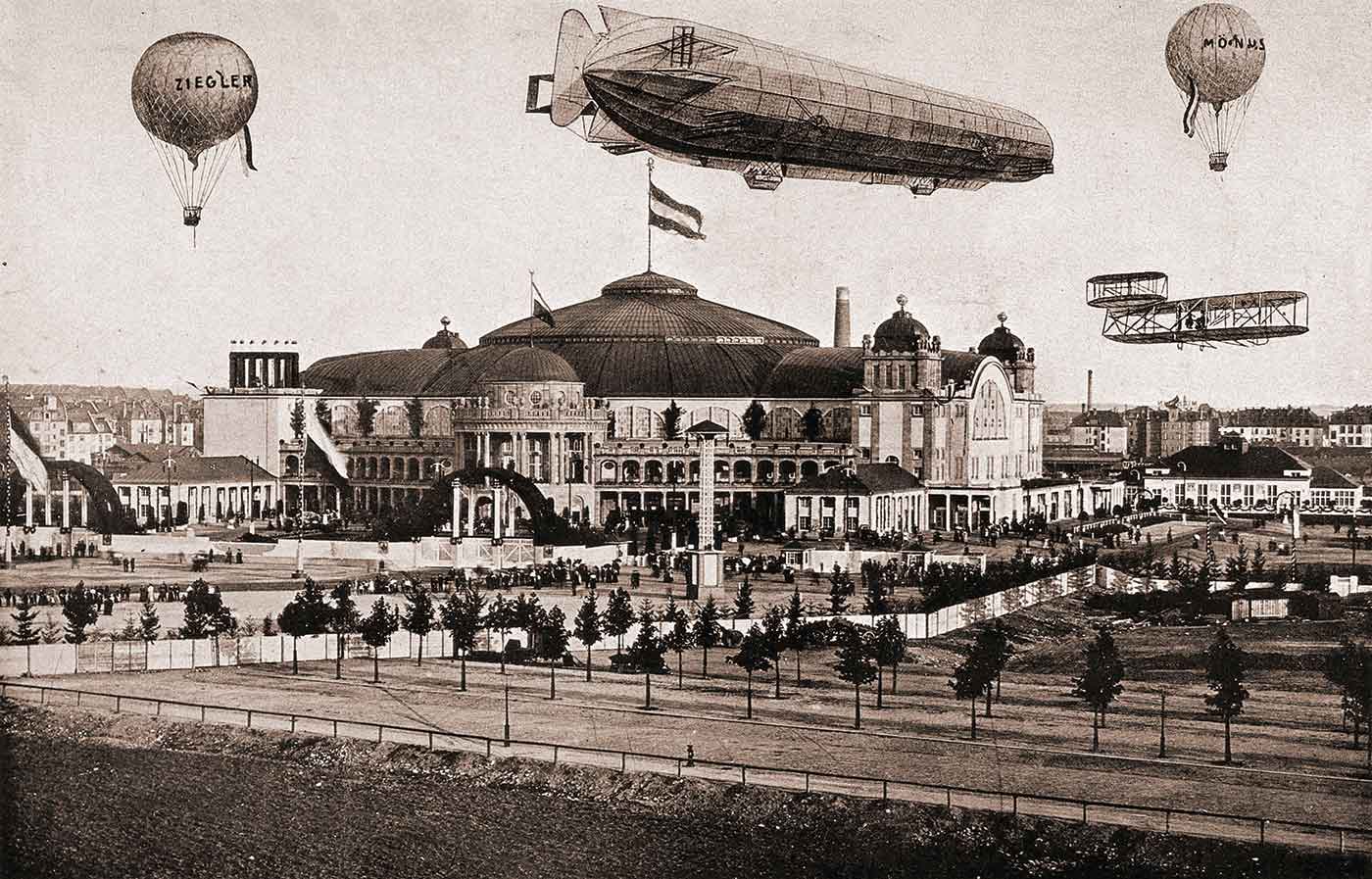 A large air-ship, aeroplane and hot-air balloons fly over a building and its grounds.