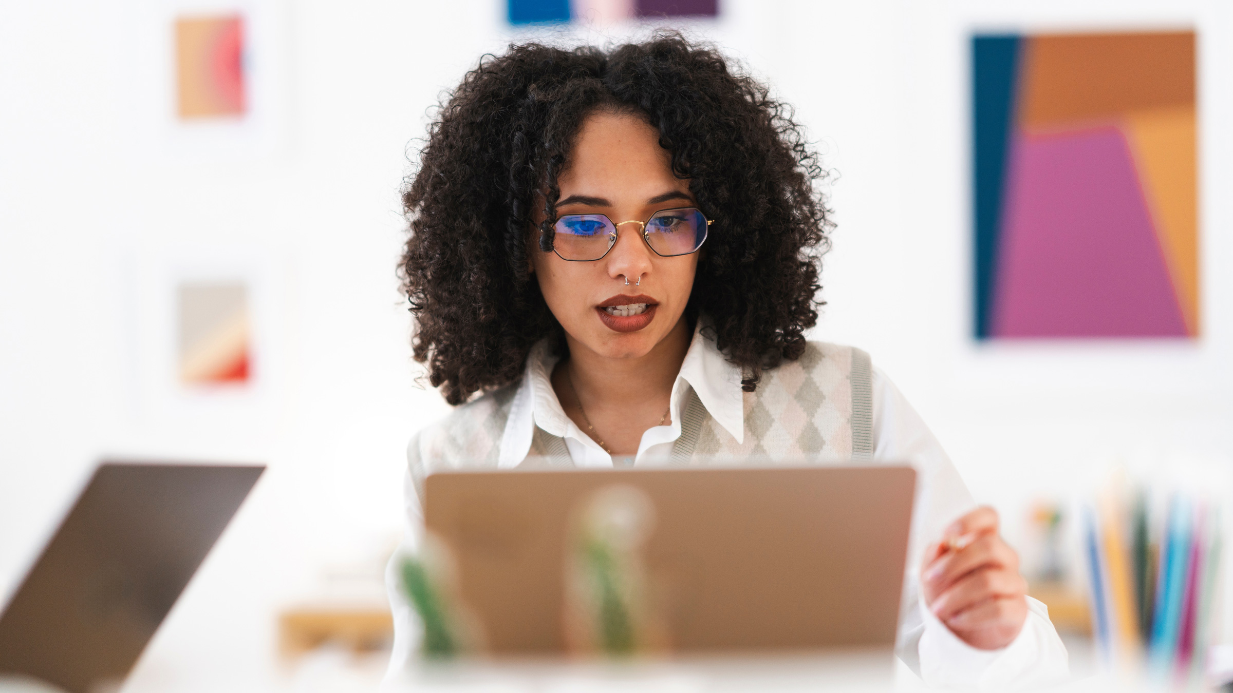 A young woman with curly hair and glasses works on her laptop in a bright, modern office space with colorful abstract art on the wall
